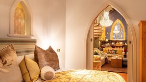 A bedroom with a large bed, a chandelier, and a view into a dining area through an arched doorway.