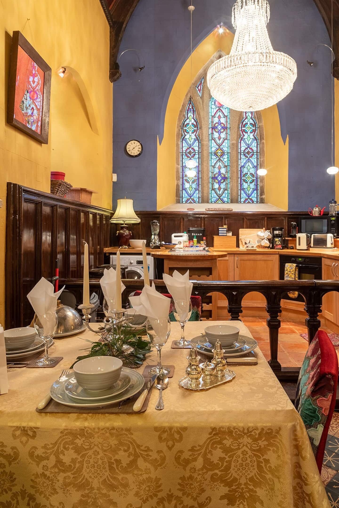 Un comedor con una mesa dispuesta para una comida, una araña, vidrieras y una cocina al fondo.