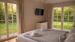 Un dormitorio con una cama, un televisor montado en la pared y ventanas grandes con cortinas.