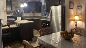 A kitchen with dark wooden cabinets, a stainless steel refrigerator, a dining table with a lace tablecloth, and a bowl of fruit.
