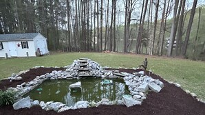 A pond with a stone border and a small waterfall, surrounded by a grassy area and trees.