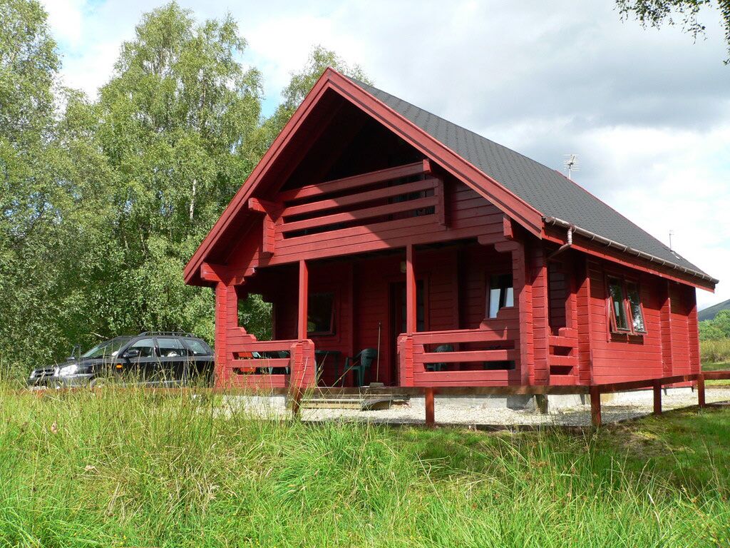 Une cabane en bois rouge, avec un toit en pente, une véranda et une voiture garée à proximité.