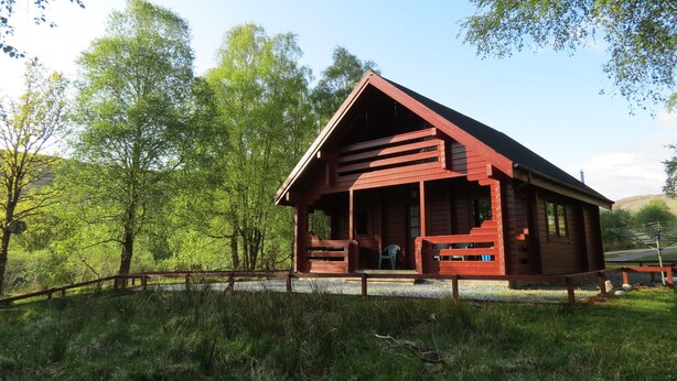 En stuga av trä med en veranda och en överbyggd entré, omgiven av frodig grönska och träd.