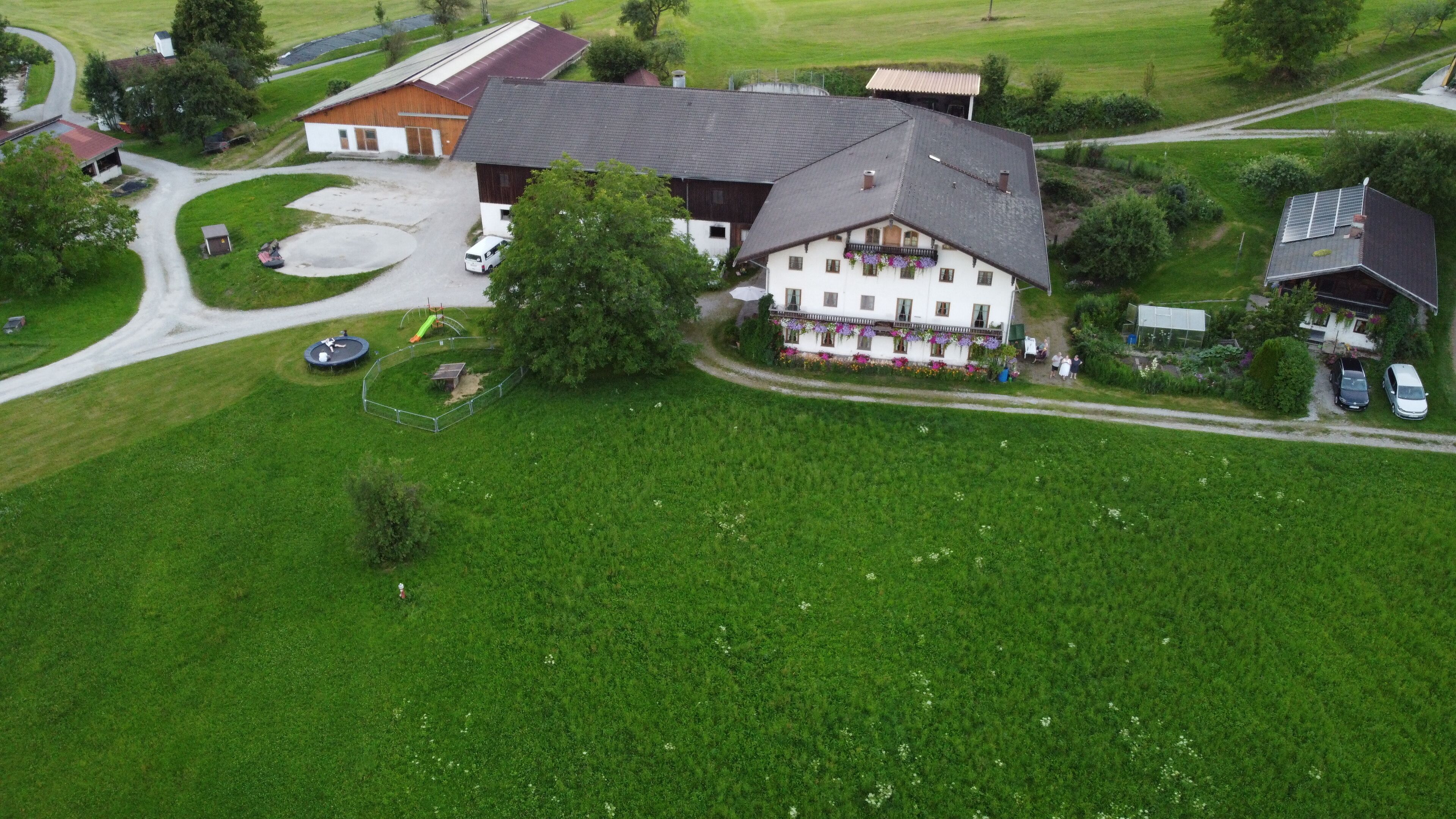 An aerial view of a large, two-story house with a sloped roof, surrounded by green fields and a winding road.