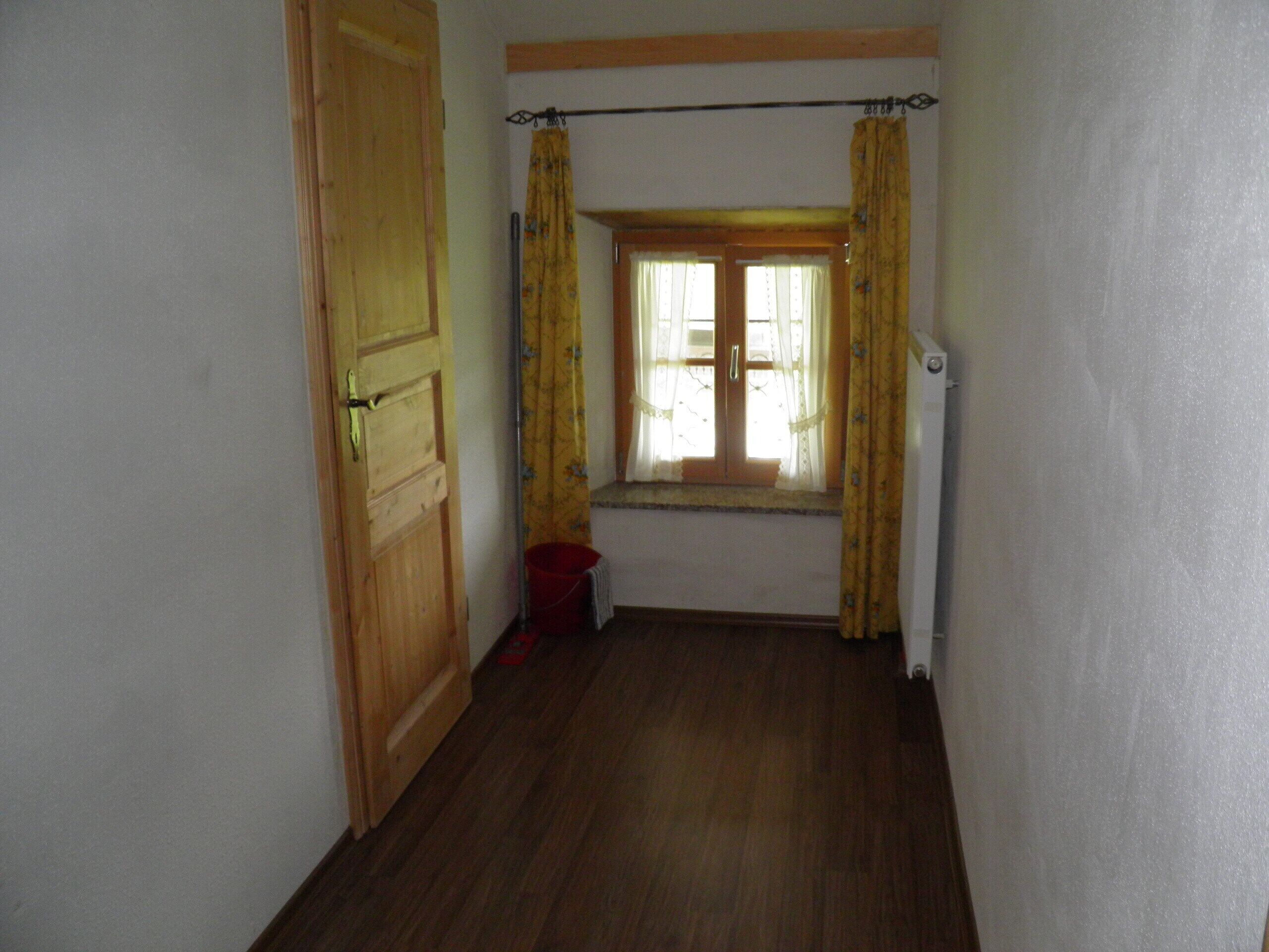 A narrow hallway with wooden flooring, a wooden door on the left, and a window with yellow curtains.