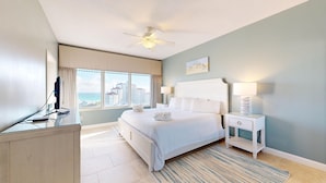 A hotel room with a bed, a desk, a chair, a lamp, a ceiling fan, and a window with a view of buildings and the sea. - Tops'l Summit 812 ~ Gulf views and quick walk to the beach (Miramar Beach)