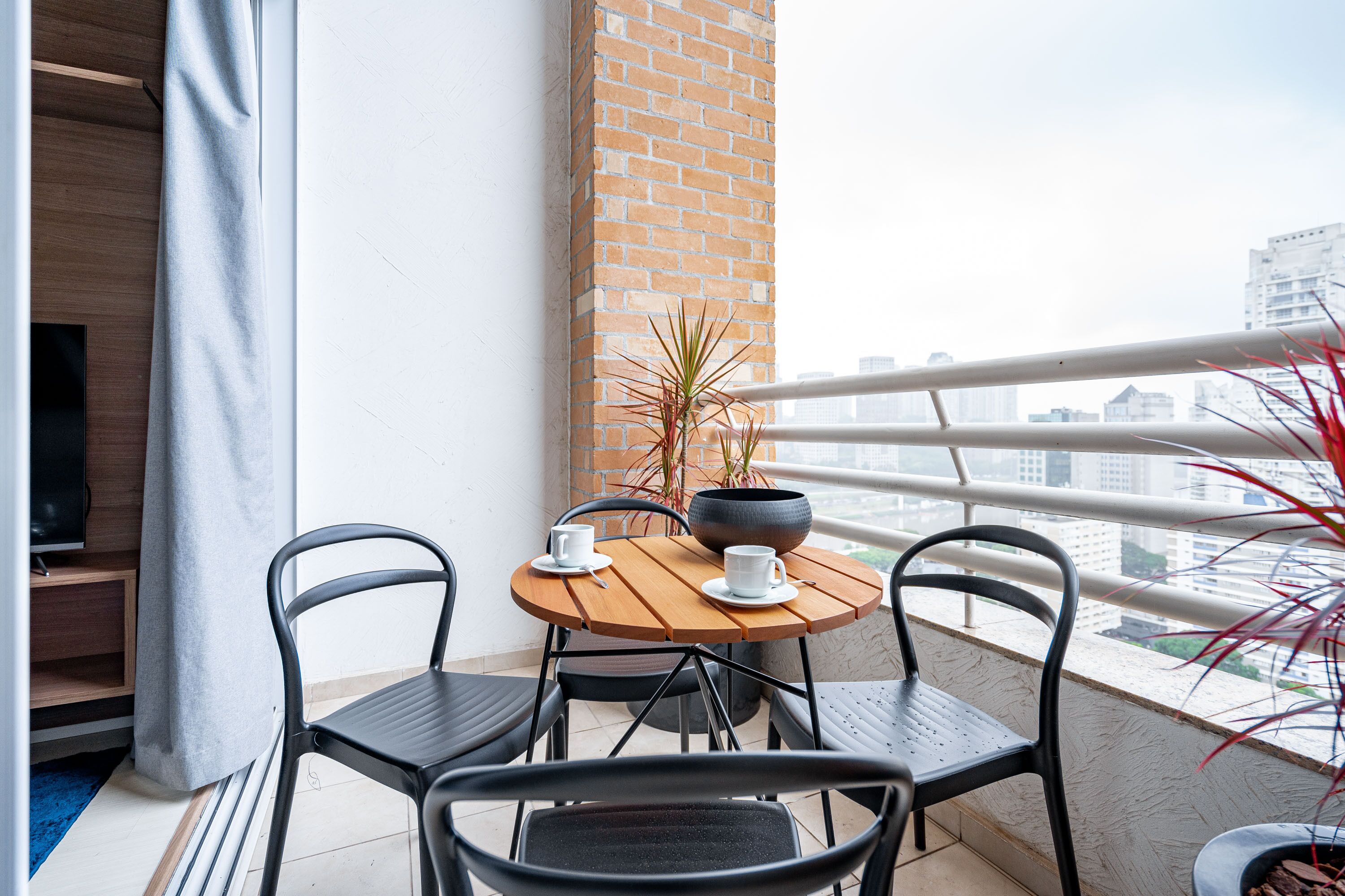A balcony with a wooden table, two chairs, a potted plant, and a view of a cityscape.