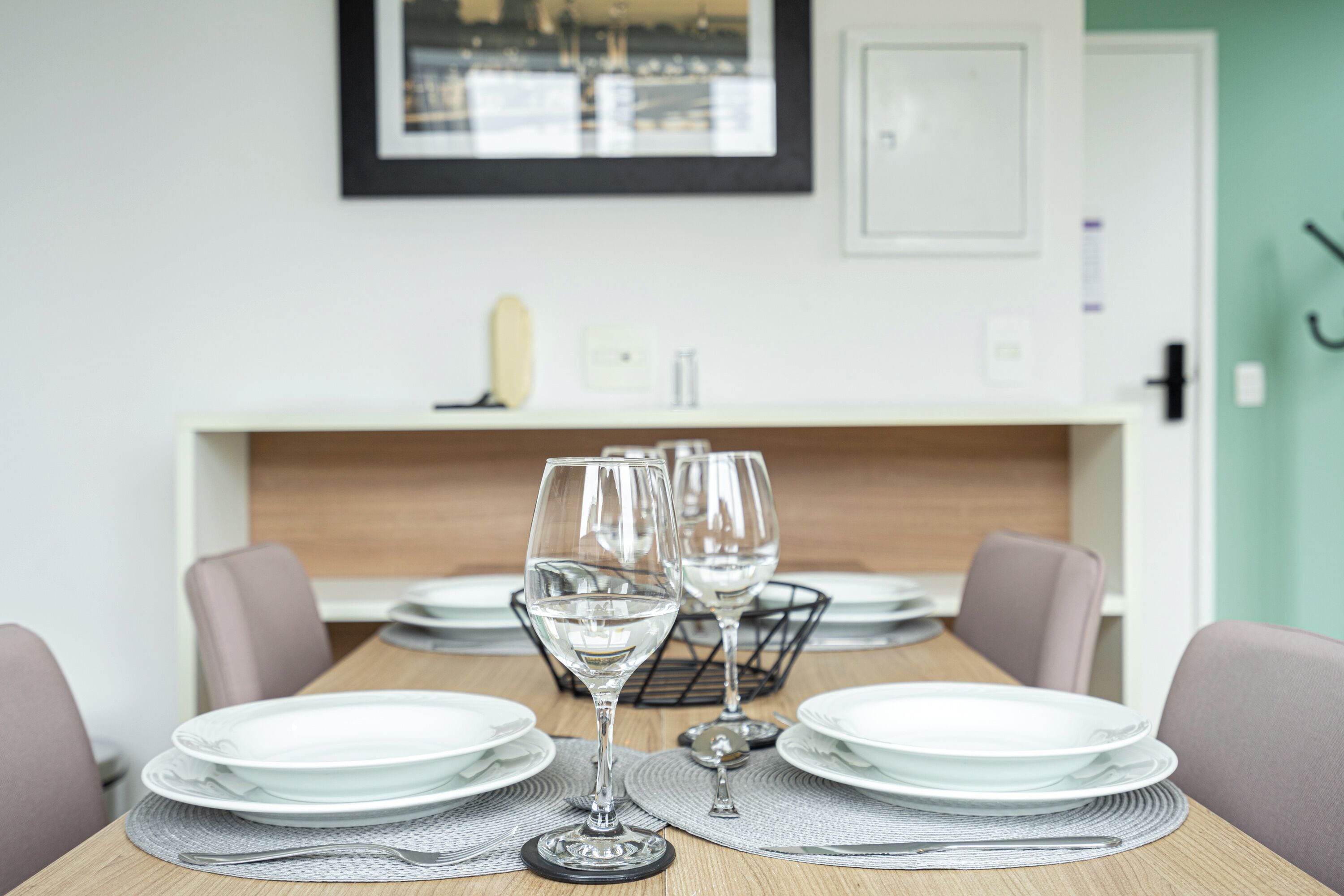Una mesa de comedor dispuesta con platos blancos y vasos transparentes, un cuadro en la pared y una pared de color claro.
