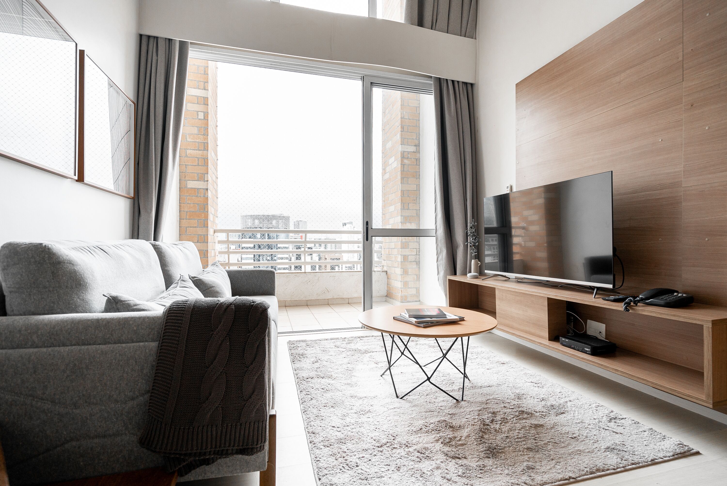 A modern living room with a grey sofa, a coffee table, a flat-screen TV on a wooden stand, and a large window with a view of buildings.