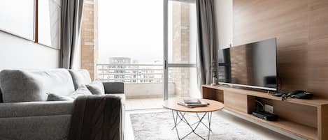 A modern living room with a grey sofa, a coffee table, a flat-screen TV on a wooden stand, and a large window with a view of buildings.