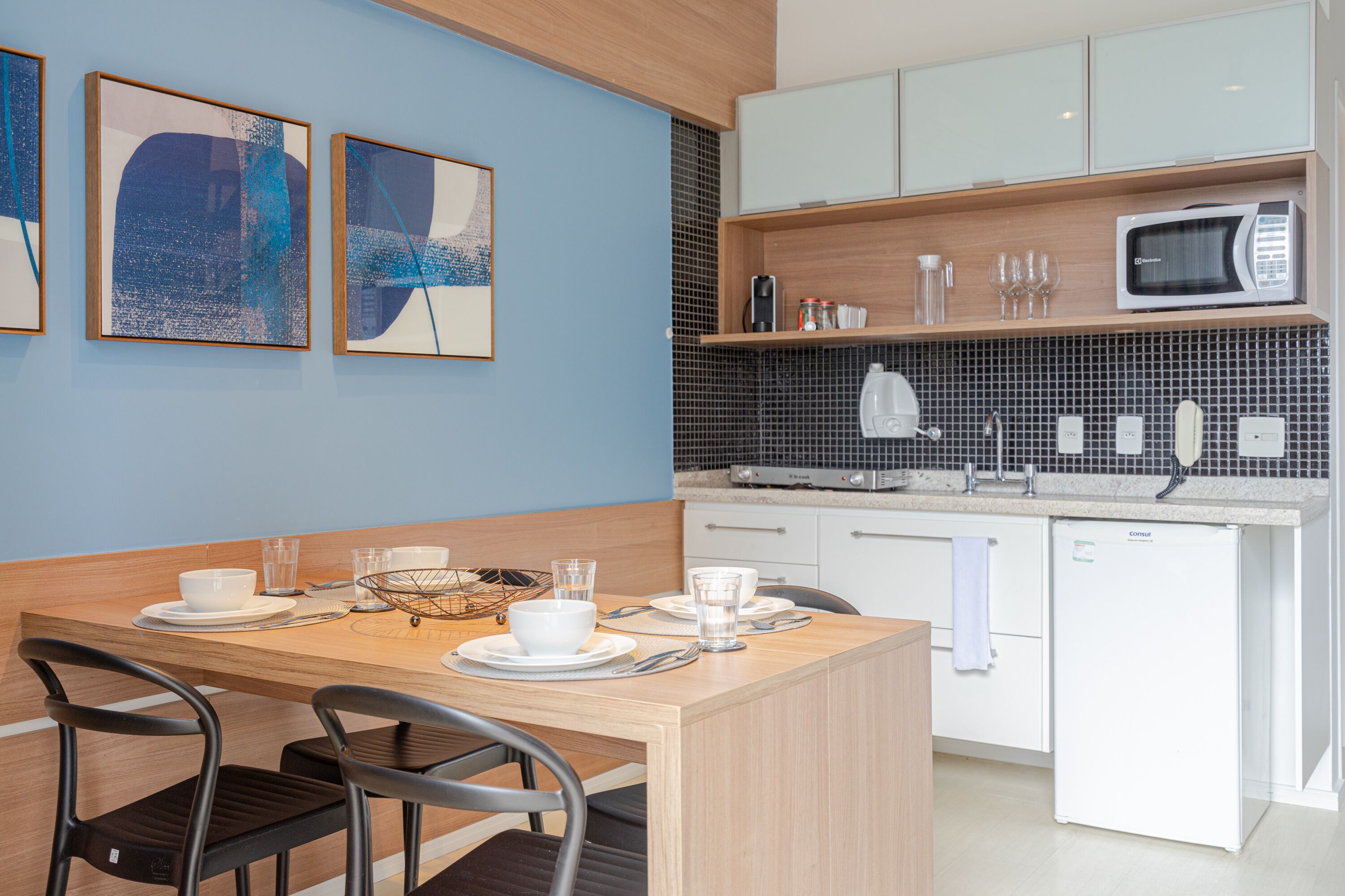 A modern kitchen with white cabinets, a microwave, and a coffee maker. There is a dining table with chairs and a small island with a bowl and glasses.