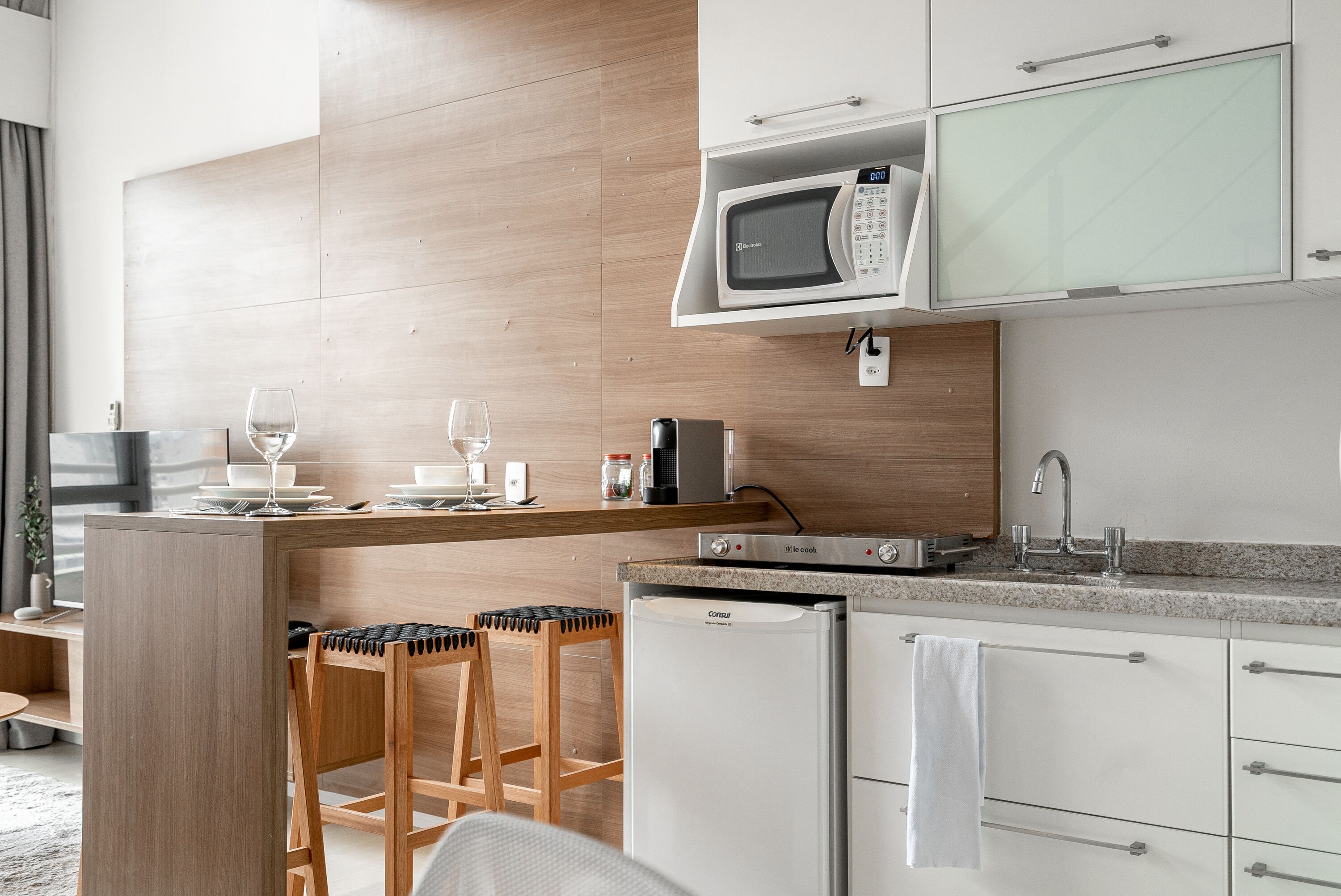 A modern kitchen with a microwave, coffee maker, and refrigerator. There are two bar stools and a small table with wine glasses.