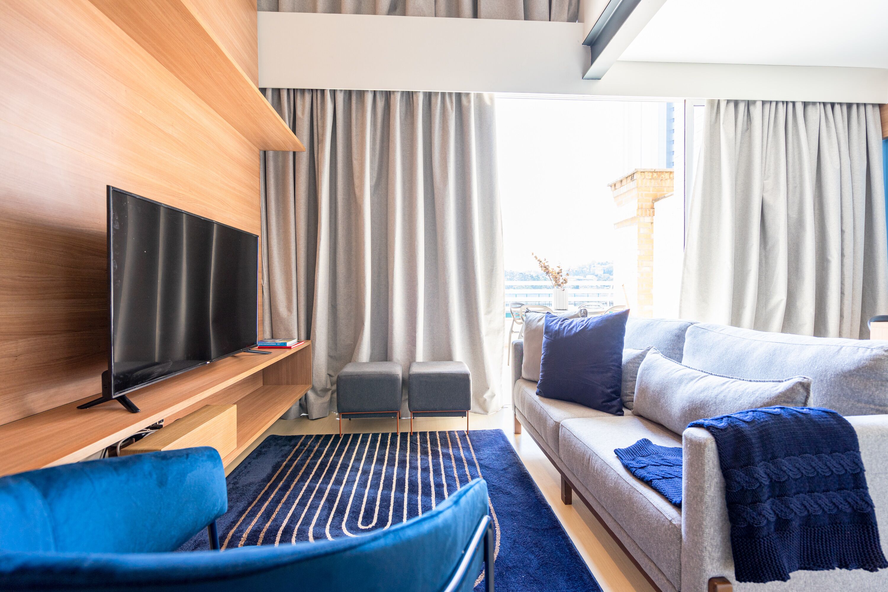 Een moderne hotelkamer met een flatscreen televisie, een bank, een blauwe fauteuil en een groot raam met gordijnen.