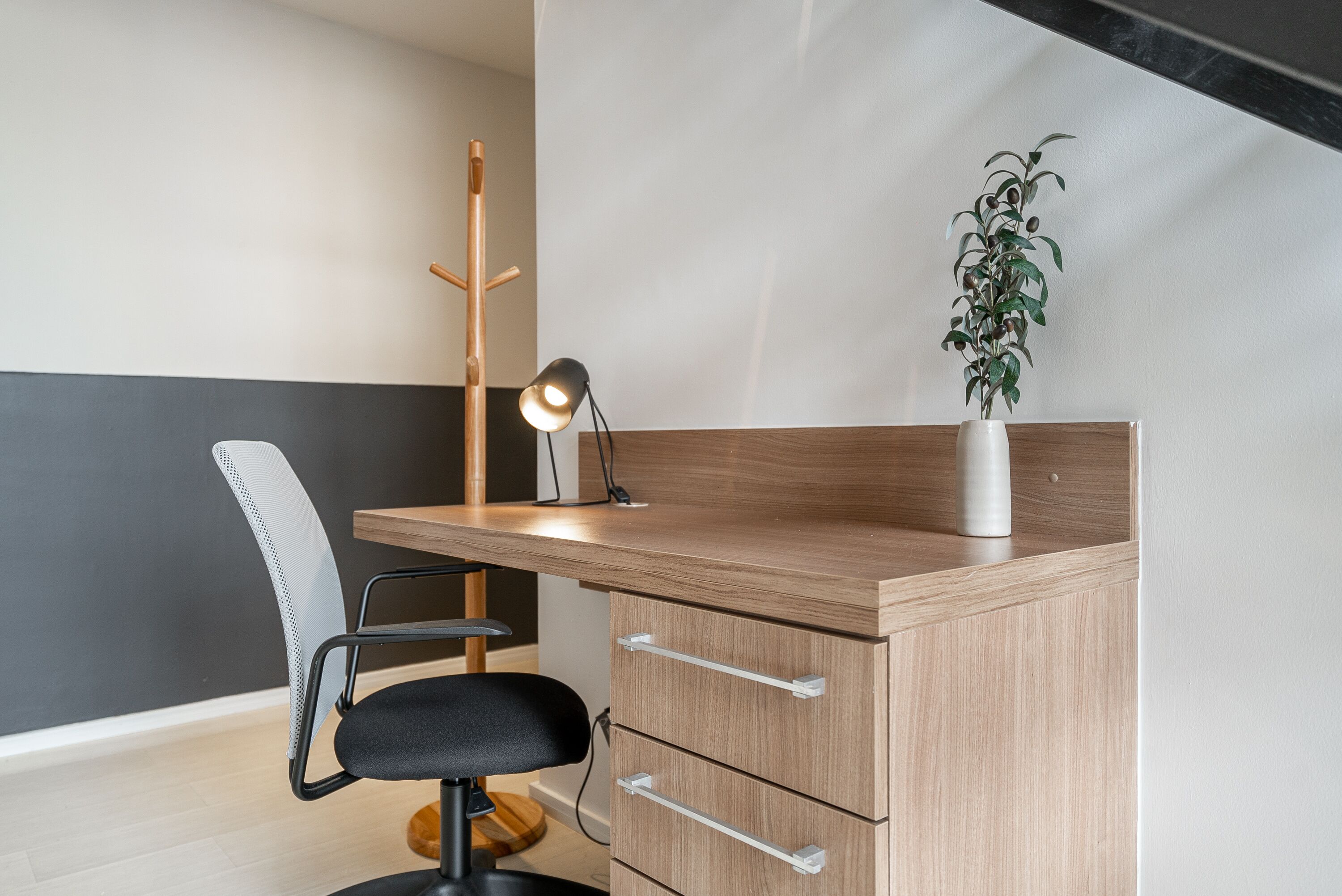 A modern office desk with a wooden finish, a black office chair, a desk lamp, a potted plant, and a coat rack.