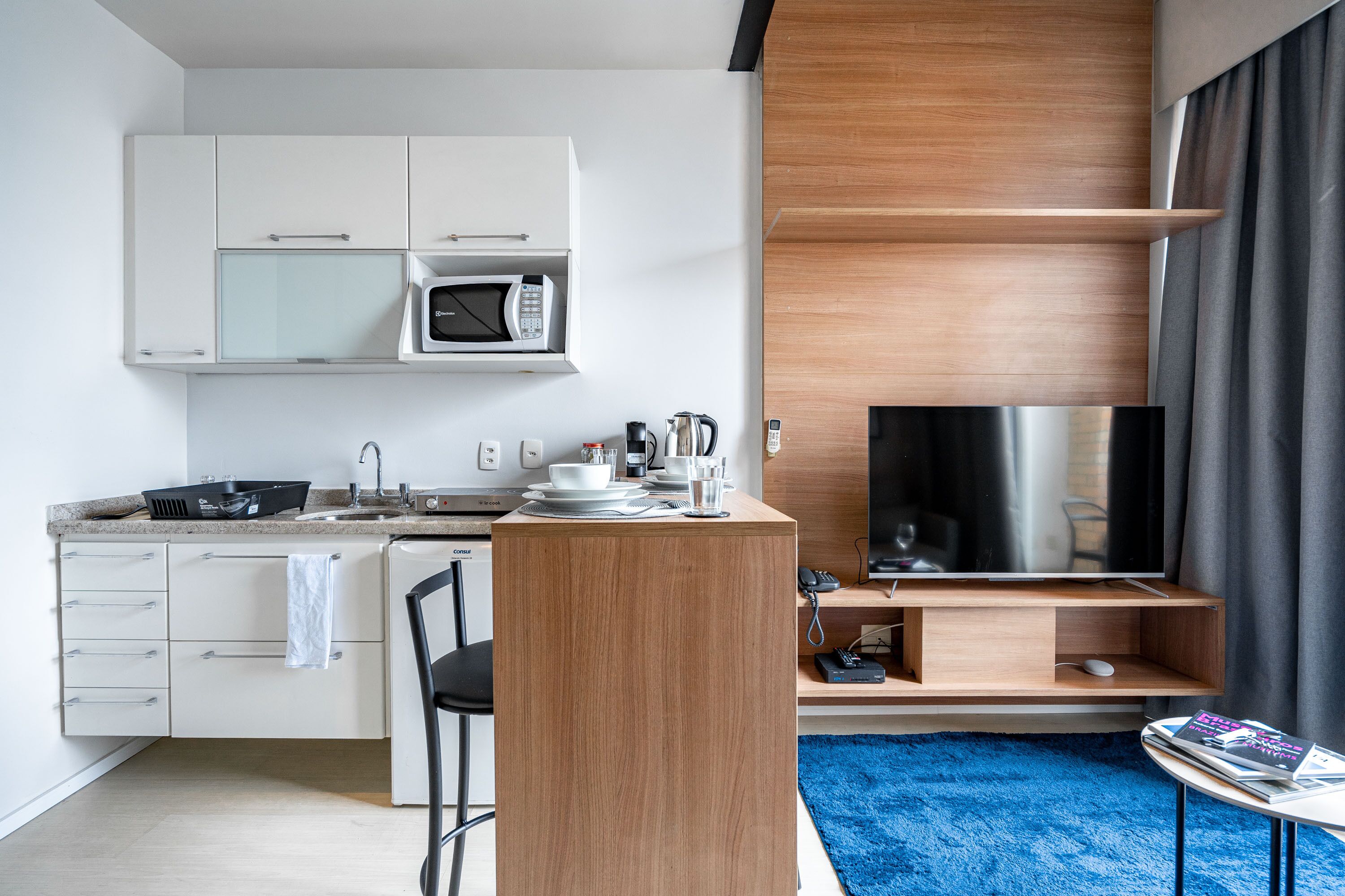 A modern kitchen with white cabinets, a microwave, and a coffee maker. There's a wooden counter with a TV and a small table with magazines.