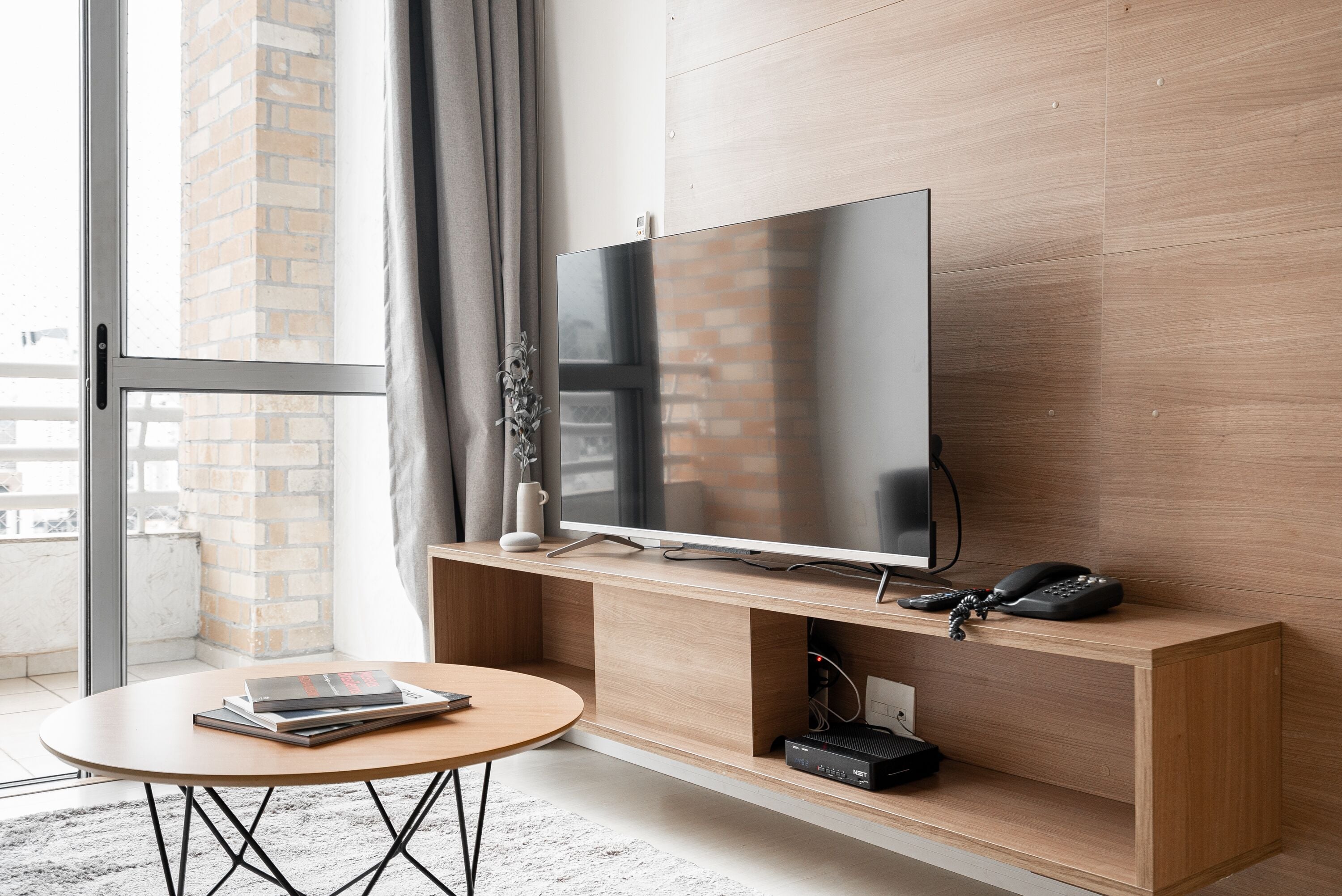 Un salon moderne avec un grand téléviseur à écran plat posé sur un meuble TV en bois, une petite table ronde avec des livres et un mur en briques en arrière-plan.