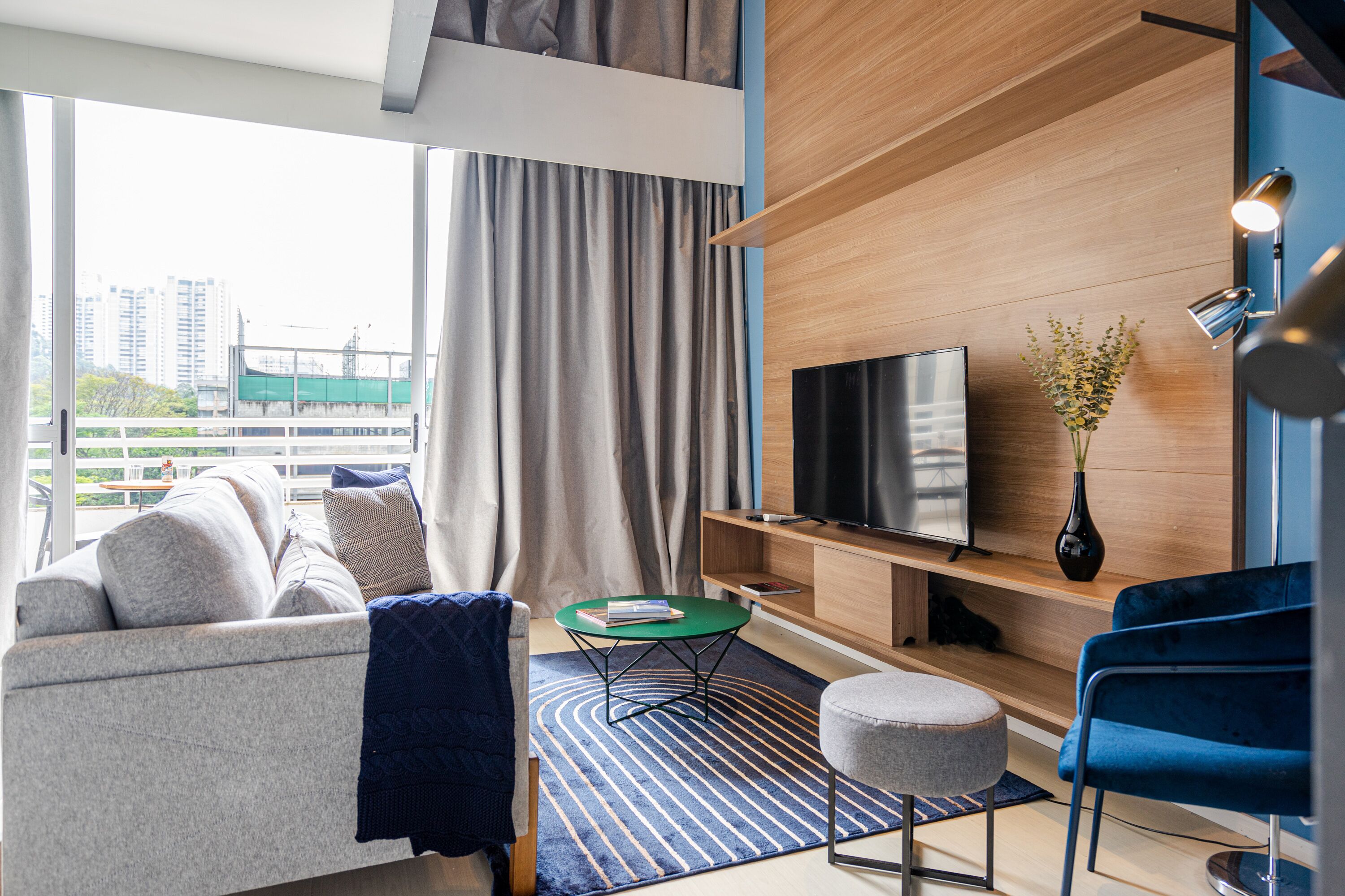 Una habitación de hotel moderna con sofá, una mesita redonda, televisor y una silla azul.