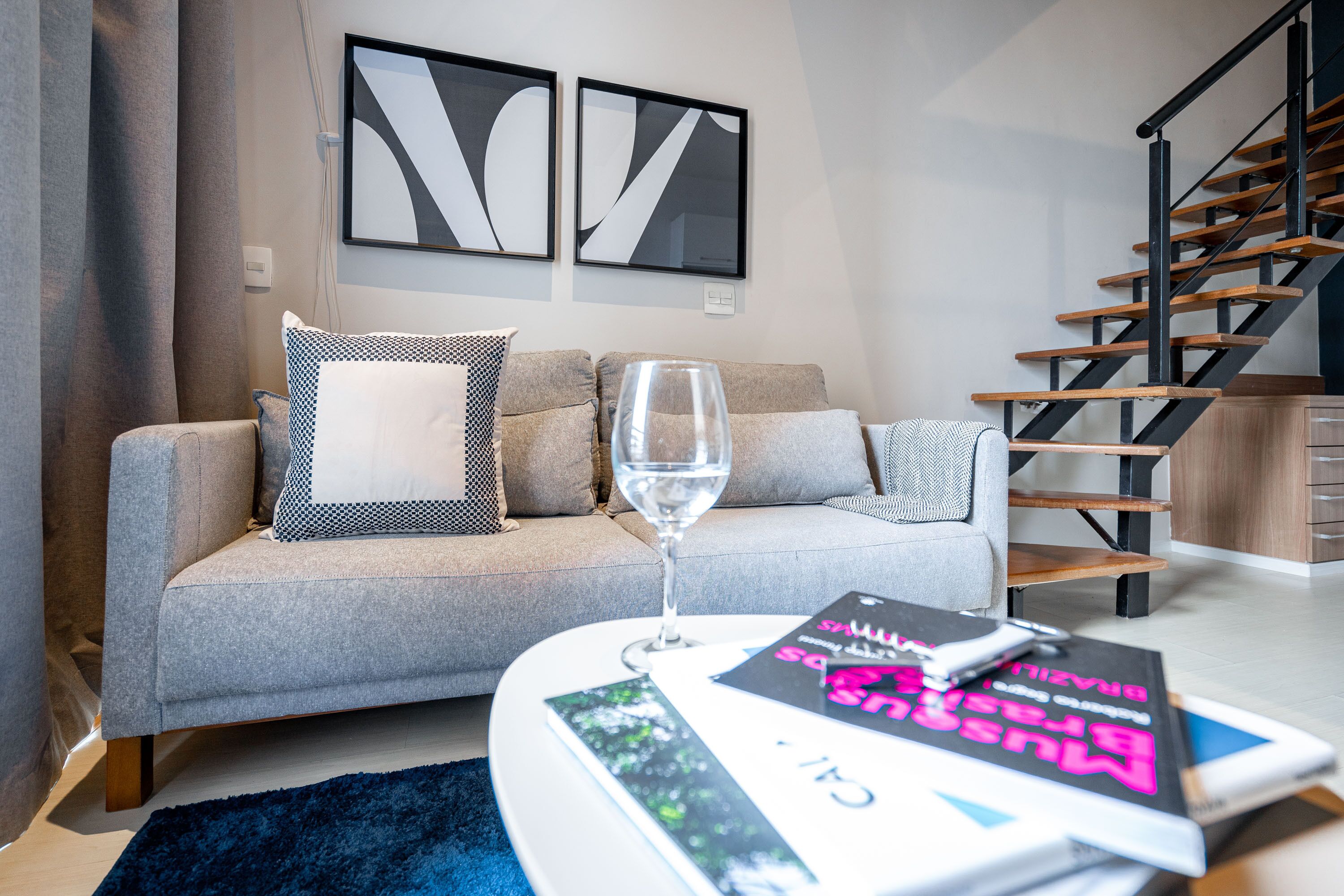 Un salon moderne avec un canapé gris, une table basse en verre et un livre. On aperçoit, à l’arrière-plan, un escalier avec des marches en bois et une rampe noire.