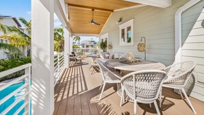 Eine überdachte Terrasse im Freien mit Holzboden, weißen Korbmöbeln und Blick auf das Meer und Palmen.