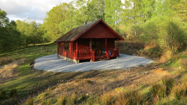A small, single-story cabin with a porch, surrounded by a gravel driveway and lush greenery. - Oak Lodge Cosy Cabin With 2 Bedrooms. Open all year. 15 miles from Fort William (Roybridge)