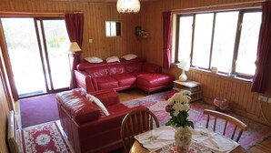 A cozy room with a red leather sofa, a wooden dining table with floral placemats, and a large window with curtains. - Oak Lodge Cosy Cabin With 2 Bedrooms. Open all year. 15 miles from Fort William (Roybridge)