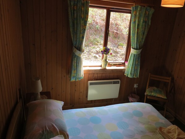 A wooden room with a bed, a window with curtains, a radiator, and a chair.