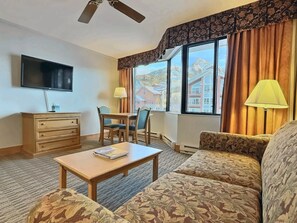 A hotel room with a flat-screen TV, a wooden desk with two chairs, a patterned sofa, a small table with a book, and a window with curtains.
