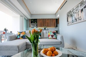 A modern living room with a sofa, colorful cushions, a glass coffee table with oranges, and a kitchen in the background.