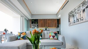 Un salon moderne avec un canapé gris, des coussins aux motifs géométriques et aux couleurs vives, une table basse en verre sur laquelle se trouve une assiette d’oranges, et un vase rempli de tulipes.