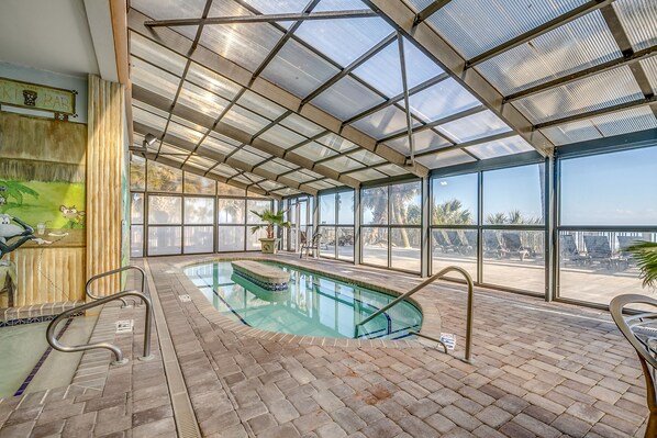 An indoor swimming pool with a glass ceiling, a brick floor, and a view of palm trees outside.