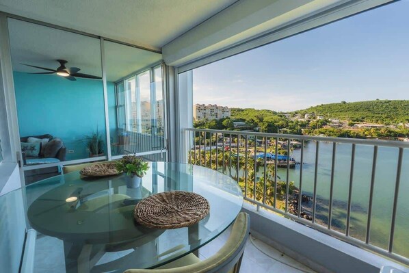 A balcony with a glass table, a ceiling fan, and a view of a river and buildings.