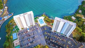 Vista aérea de una zona costera con dos edificios blancos destacados, un puerto deportivo con numerosos barcos y un estacionamiento con varios coches.