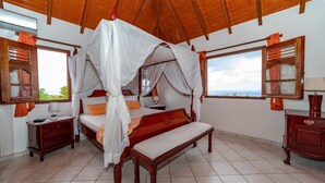 Una cama con dosel sobre cuatro postes, cabecera de madera, dos mesitas de noche y vista al mar a través de un ventanal grande.
