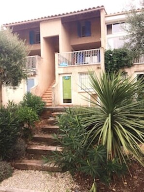 Set amidst lush greenery, this two-story building features a balcony and a flat roof, with a charming entrance accessible via outdoor stairs.