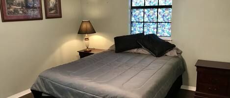 Un dormitorio con cama, mesita de noche con lámpara, cómoda y ventana con vidrieras.