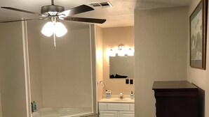 A bedroom with a bed, a ceiling fan, a dark wooden cabinet, a woven basket, and a bathroom with a bathtub, sink, and lights. - Secluded Lakehouse Right on Lake Eufaula! (Crowder)