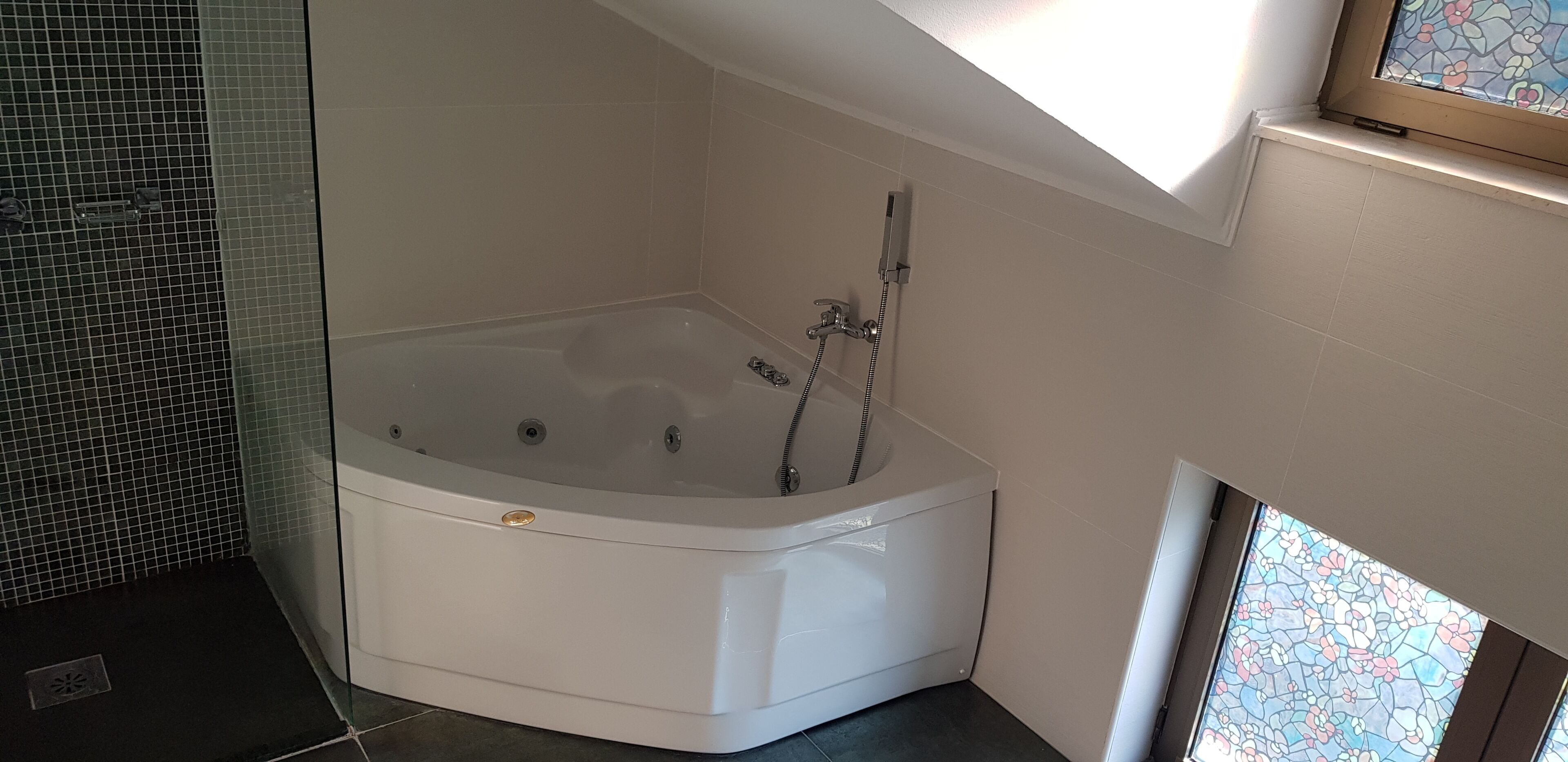 Eine Eckbadewanne mit Duschkopf, Glasabtrennung und einem Fenster mit Buntglas.
