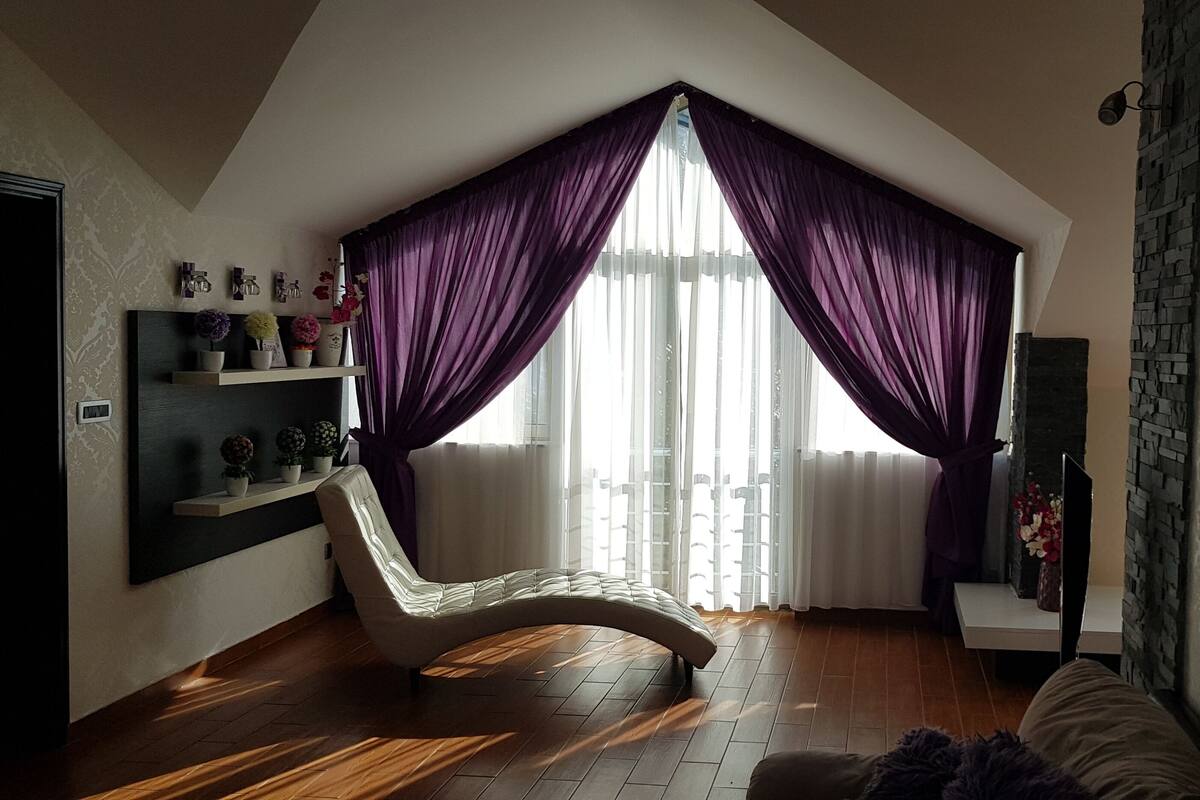 Un salon moderne avec un canapé aux lignes courbes, une grande fenêtre ornée de rideaux violets, une cheminée en pierre et une étagère murale décorative.