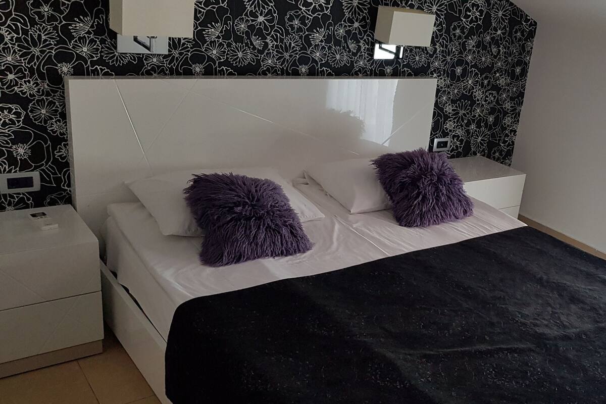 Une chambre à coucher avec une tête de lit blanche, deux coussins moelleux violets, une couvre-lit noire et un papier peint noir et blanc à motifs floraux.