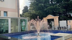 A swimming pool with a water fountain feature, surrounded by a building and trees. - Domador Rooms & Apartments (Becici)