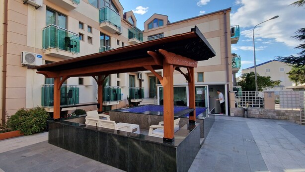 A modern outdoor seating area with a wooden pergola, a pool, and a building with balconies in the background. - Domador Rooms & Apartments (Becici)