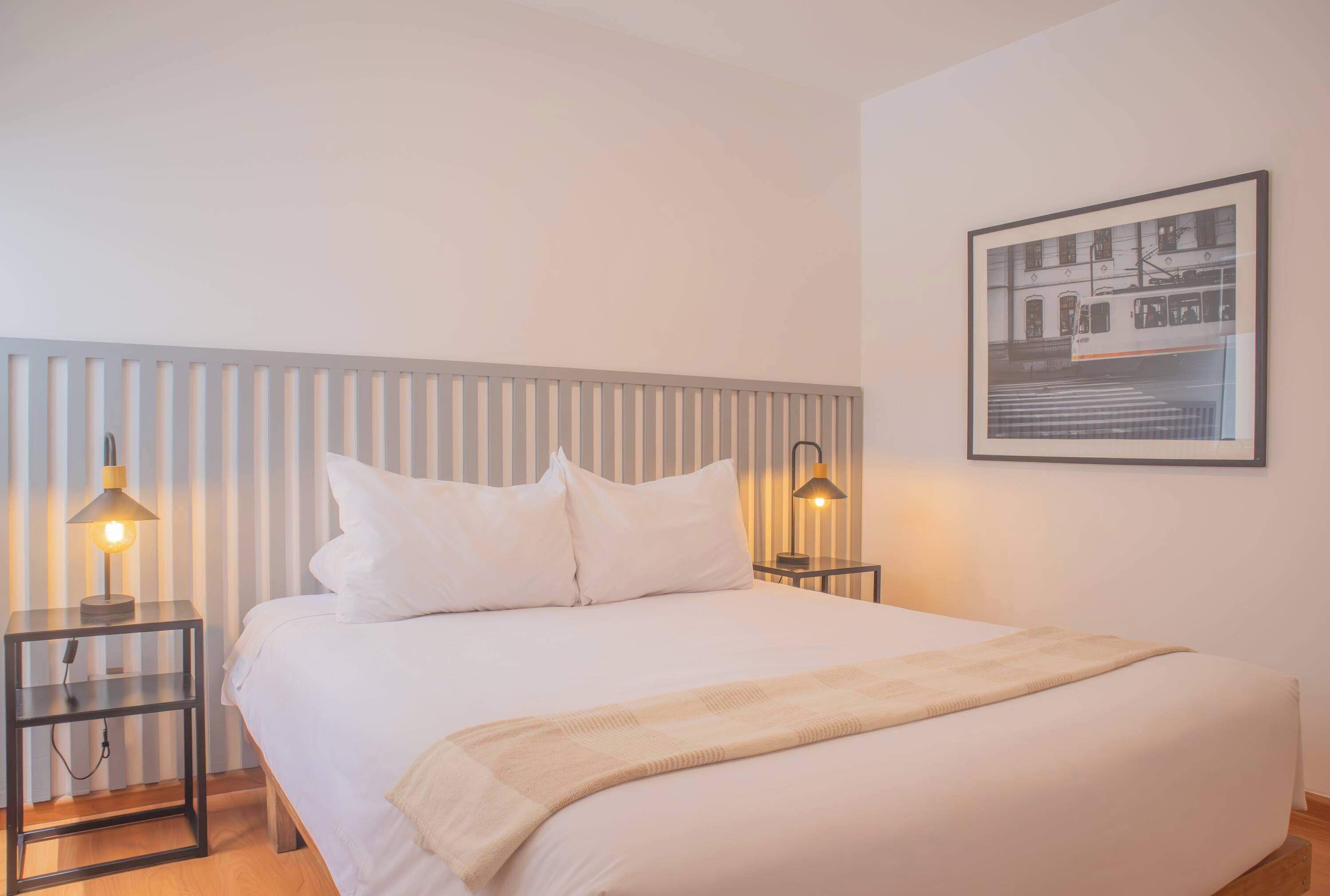 A neatly made bed with white linens and a beige throw blanket, flanked by two bedside tables with lamps, in a room with a striped headboard and a framed picture on the wall.