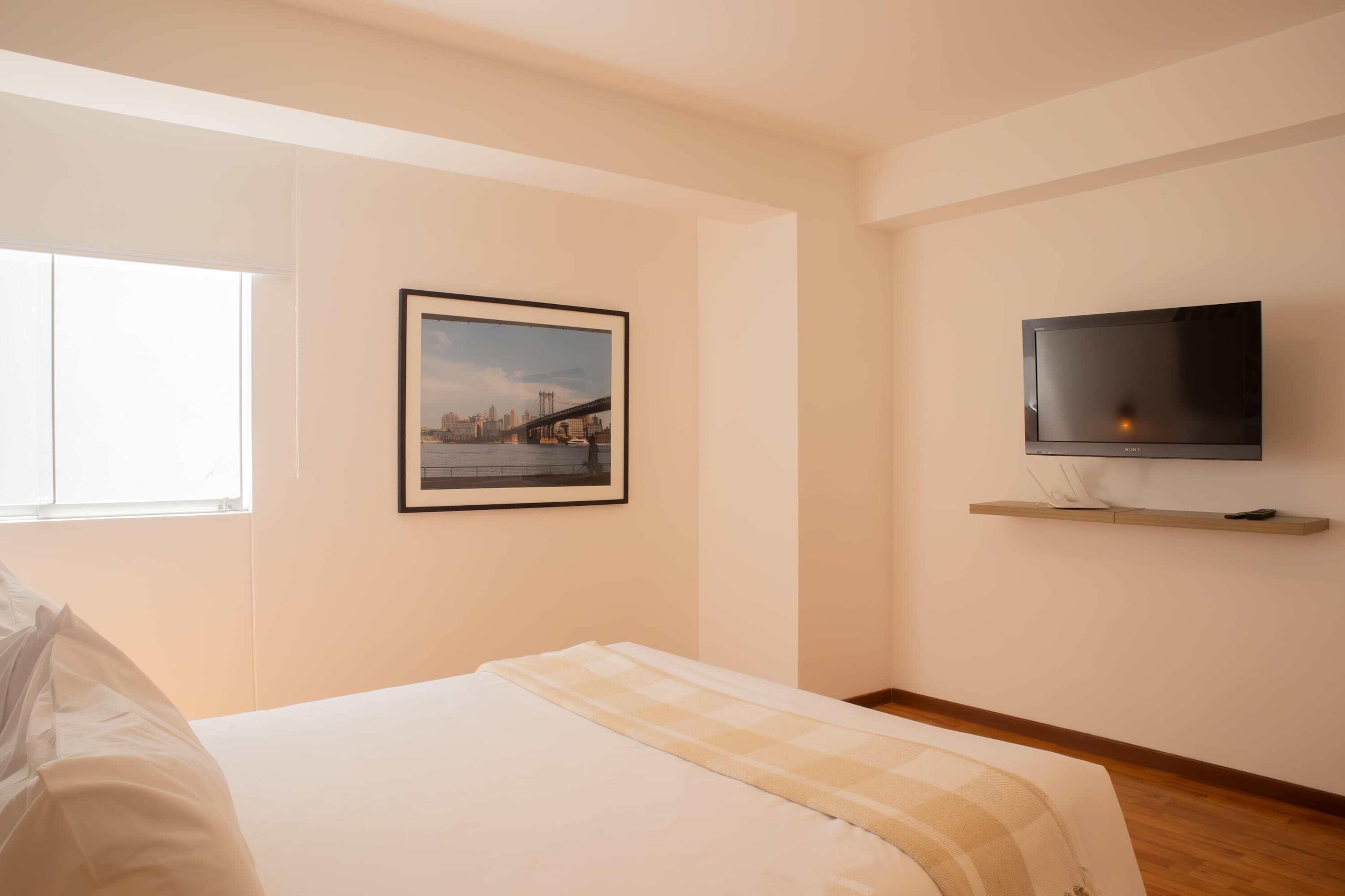 Une chambre à coucher avec un lit, un téléviseur fixé au mur, une fenêtre et un tableau encadré.