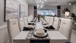 A dining area with a long table set for a meal, featuring white chairs, a modern chandelier, and artwork on the walls. - PROMO Elegant 9 BR Villa, Pool/Spa, Resort - by Blue Sky Villas (Davenport)