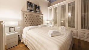 Quarto de hotel com cabeceira estofada, uma cama grande, mesa de cabeceira com luminária e guarda-roupa com cortinas.