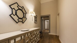 Um corredor com piso de madeira, um balcão branco com painel decorativo, uma luminária na parede e um quadro emoldurado.
