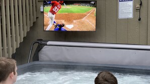 Ein Whirlpool mit zwei Personen, ein an der Wand befestigter Fernseher, der ein Baseballspiel zeigt, und ein Sicherheitshinweis an der Wand.