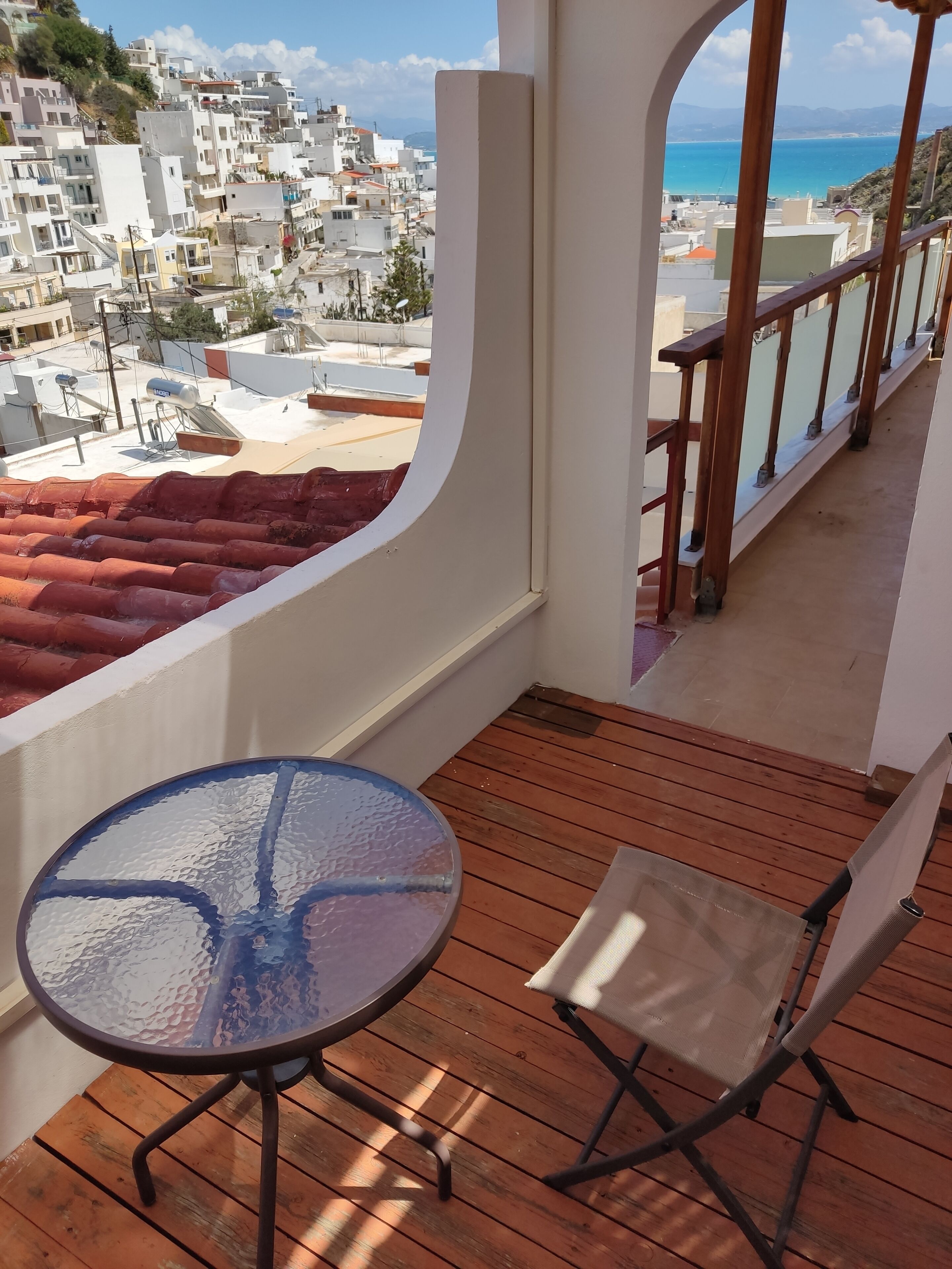 A balcony with a round glass table and a chair, overlooking a coastal town with buildings and a beach.