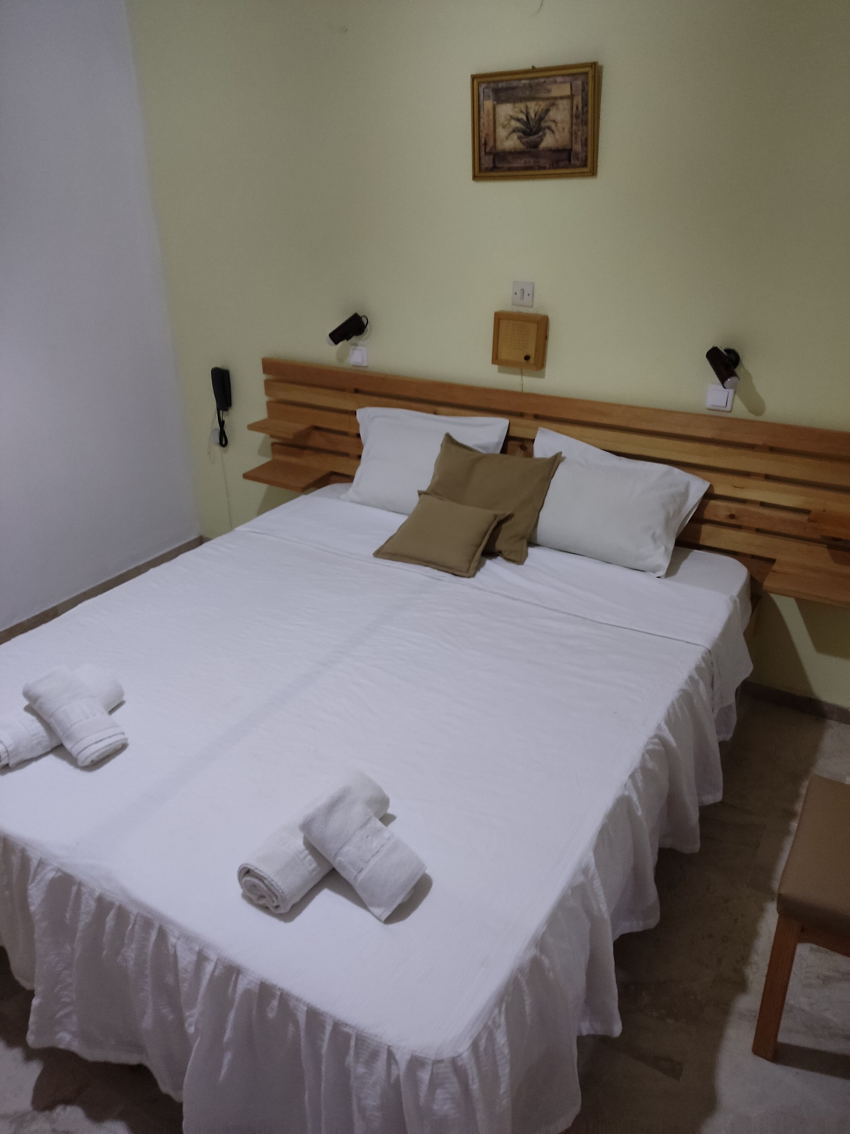 A neatly made bed with white linens and two towels, a wooden headboard, and a wall-mounted phone and light switch.