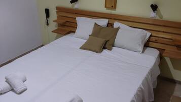 A neatly made bed with white linens and two towels, a wooden headboard, and a wall-mounted phone and light switch.